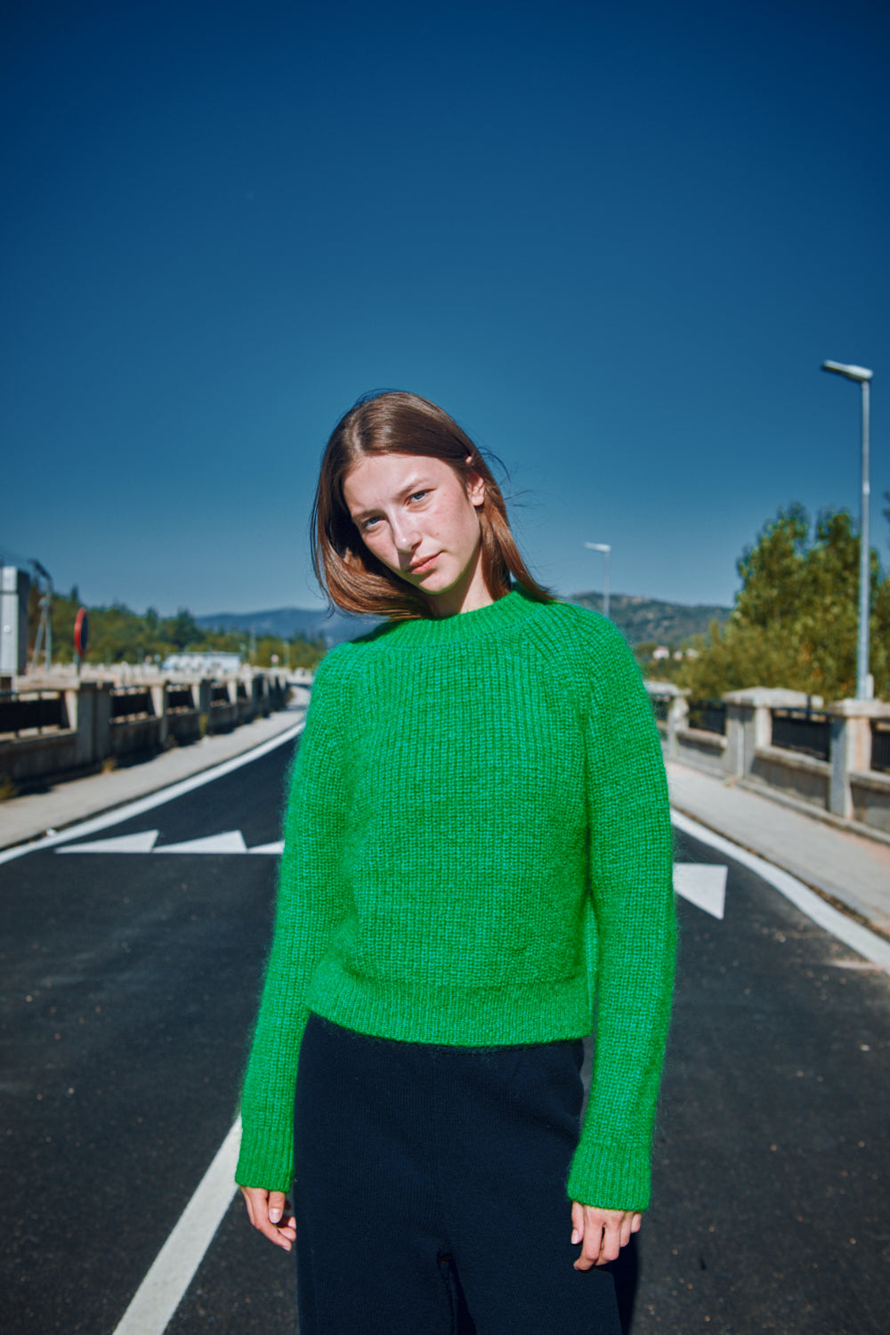 Person wearing a green sweater standing on a road with a clear blue sky.