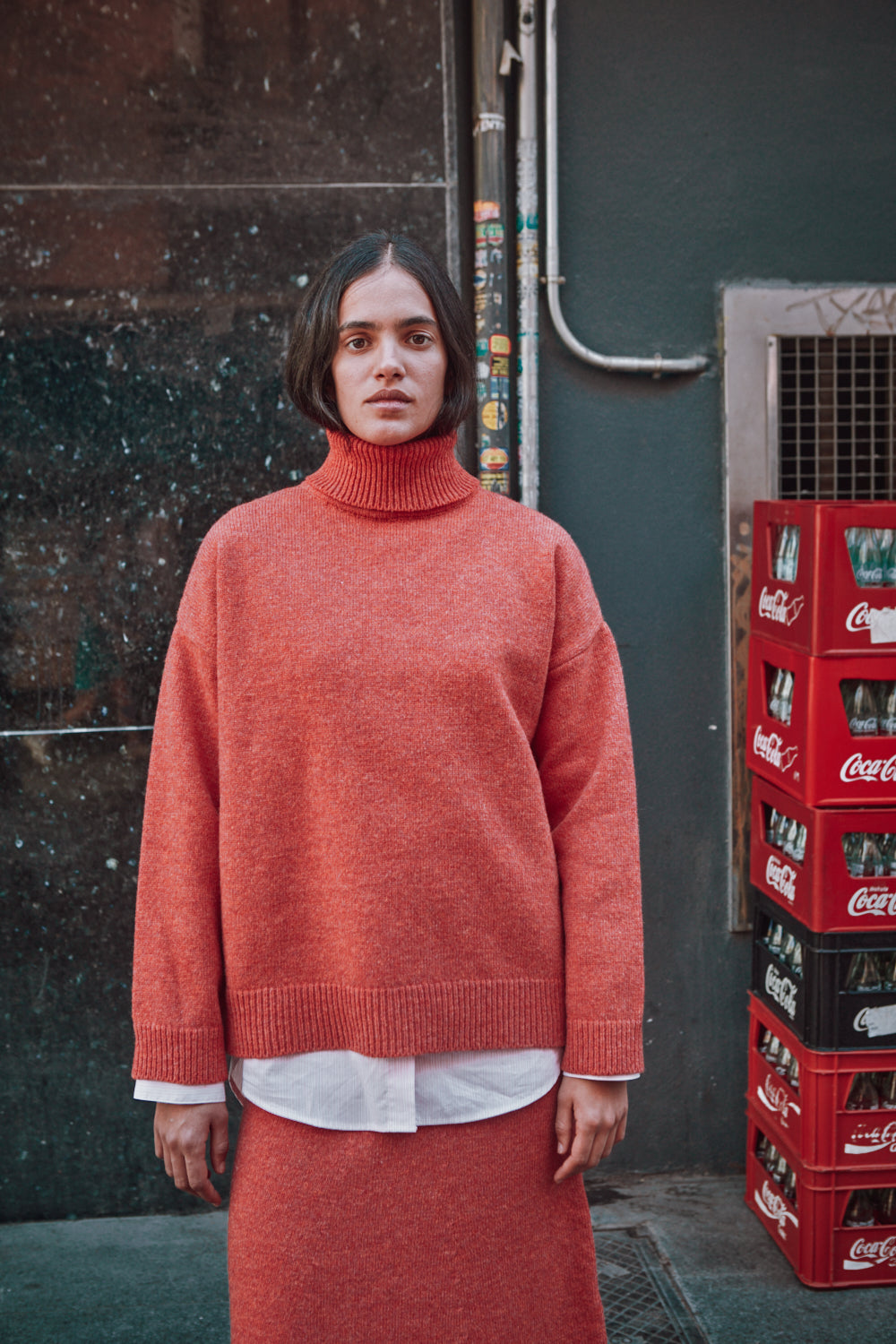 Person wearing a red L'ENVERS sweater standing next to Coca-Cola crates against a textured wall.