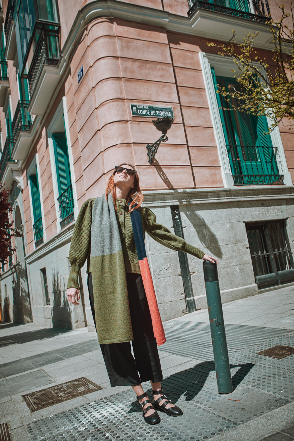 Woman standing on a street corner wearing a colorful scarf in an urban setting.