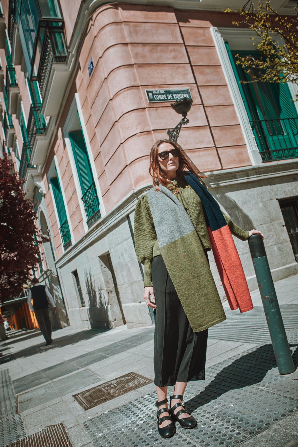 Woman standing on a street corner wearing a colorful scarf in an urban setting.