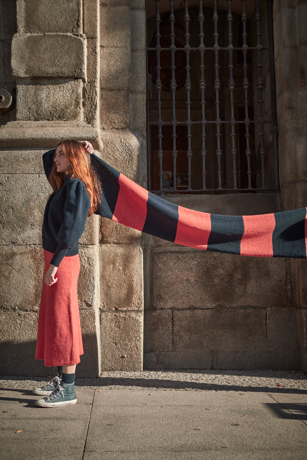 Woman wearing a long, colorful dress with a coral and navy striped scarf in front of an old building.