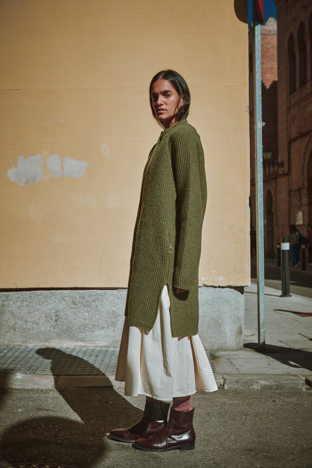 Personne portant un long pull vert et une jupe blanche au coin d'une rue.