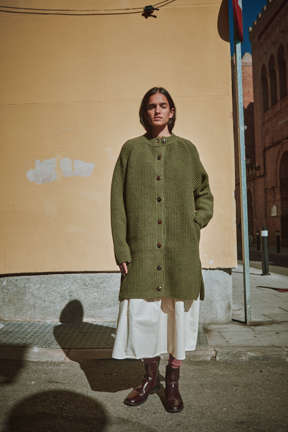 Personne portant un gilet en tricot vert, debout contre un mur beige.