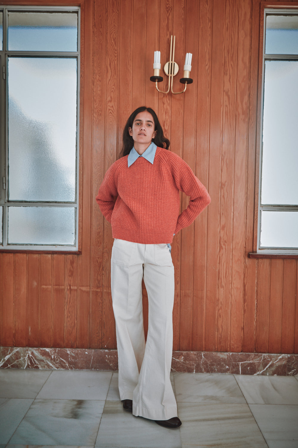 Personne portant unpull  corail  et un pantalon blanc devant un mur en bois avec un lustre.