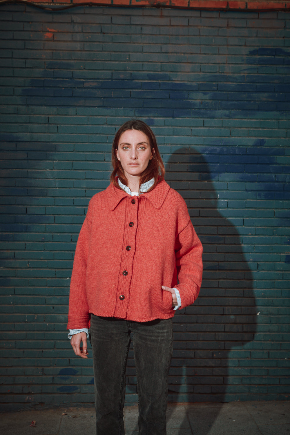 Personne portant une veste corail de L'ENVERS devant un mur texturé et une porte en bois.