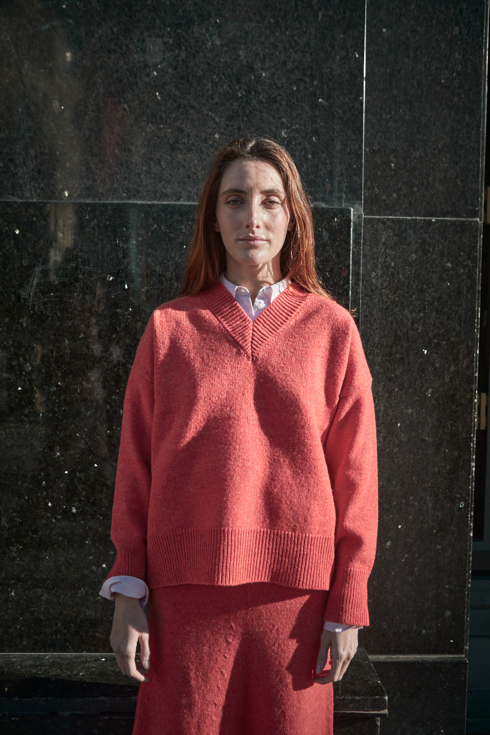 Femme portant un pull et une jupe corail, debout contre un mur de marbre foncé.