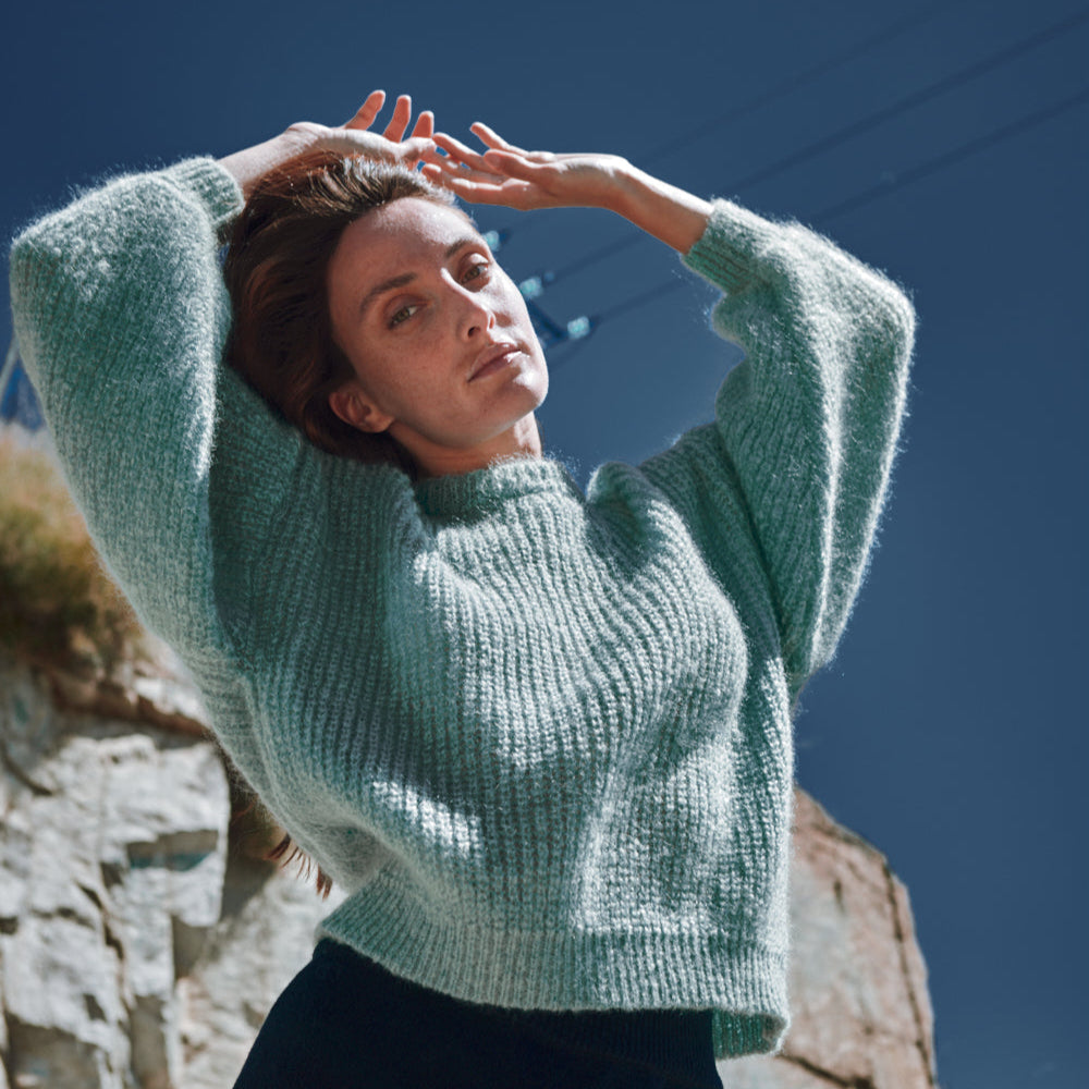 Personne portant un pull vert clair sur fond de ciel bleu et de mur de pierre. L'Envers x Marion Graux