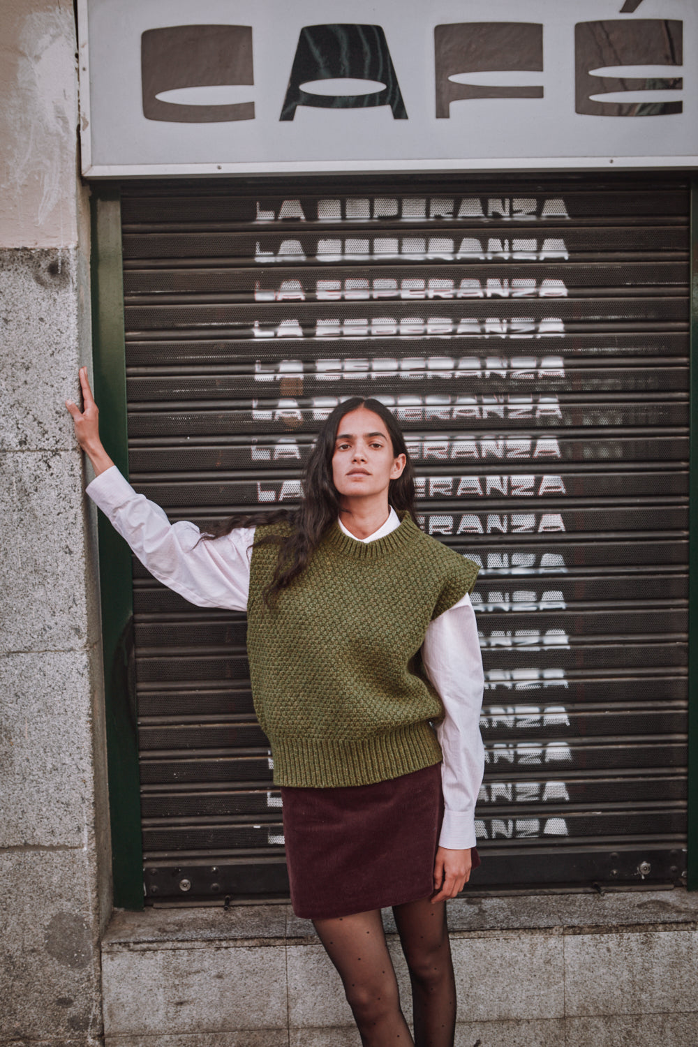 Femme debout devant un volet de café fermé, vêtue d'un pull-over vert olive.