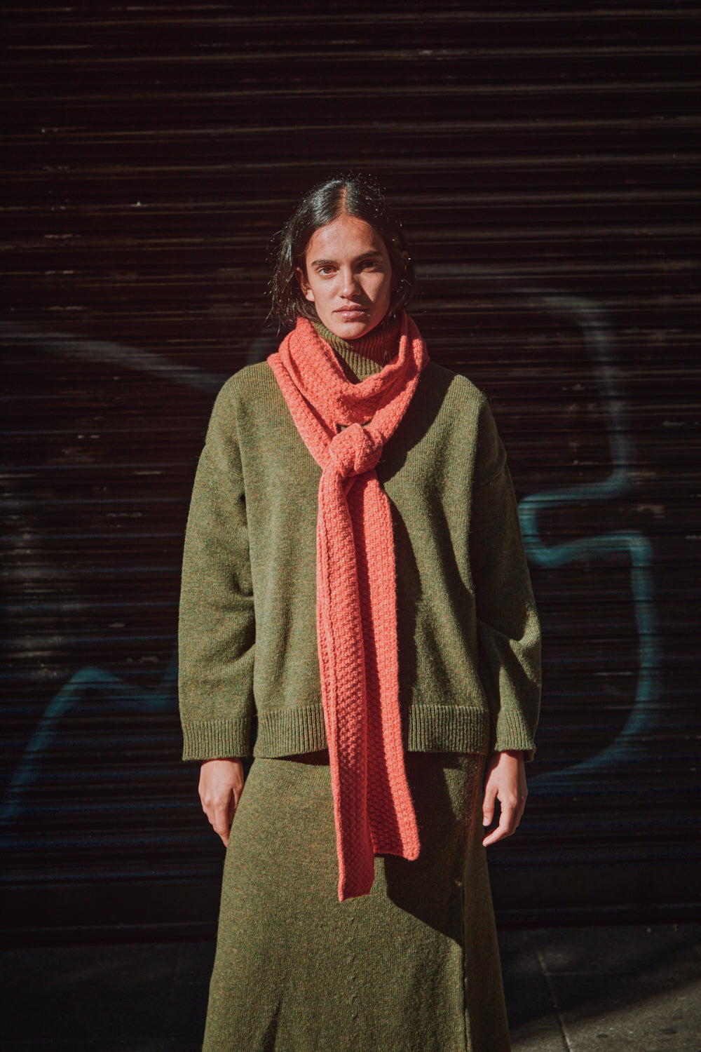 Personne portant un pull-over vert et une écharpe corail, debout sur un fond sombre