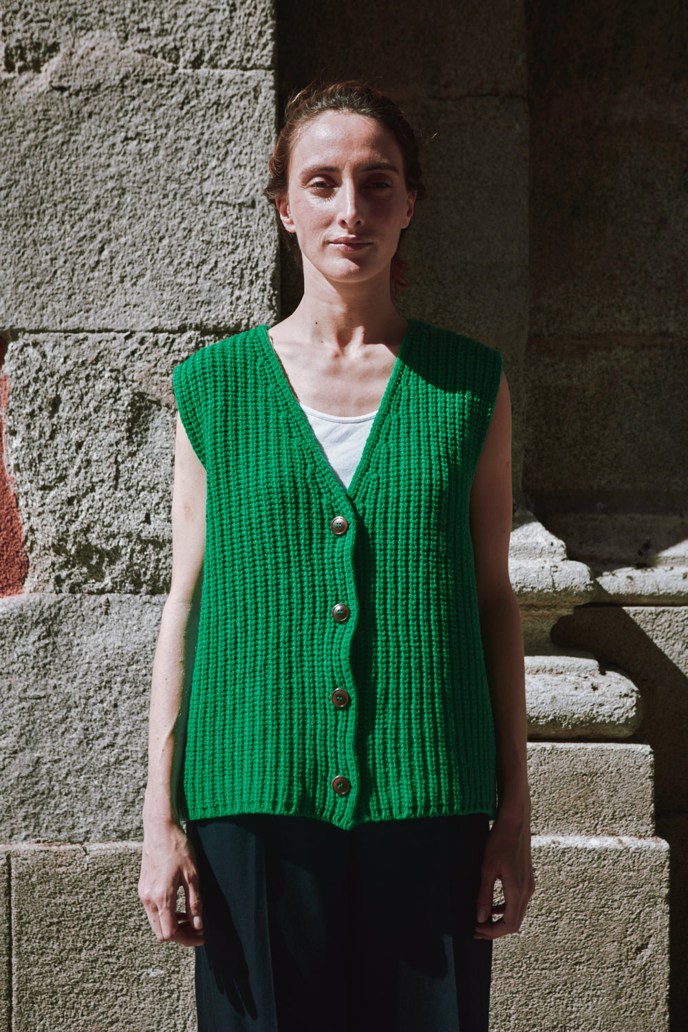 Personne portant un gilet tricoté vert, debout contre un mur de pierre.