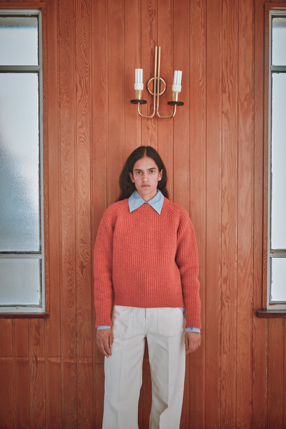 Person wearing a coral sweater and white pants standing in front of a wooden wall with a chandelier.