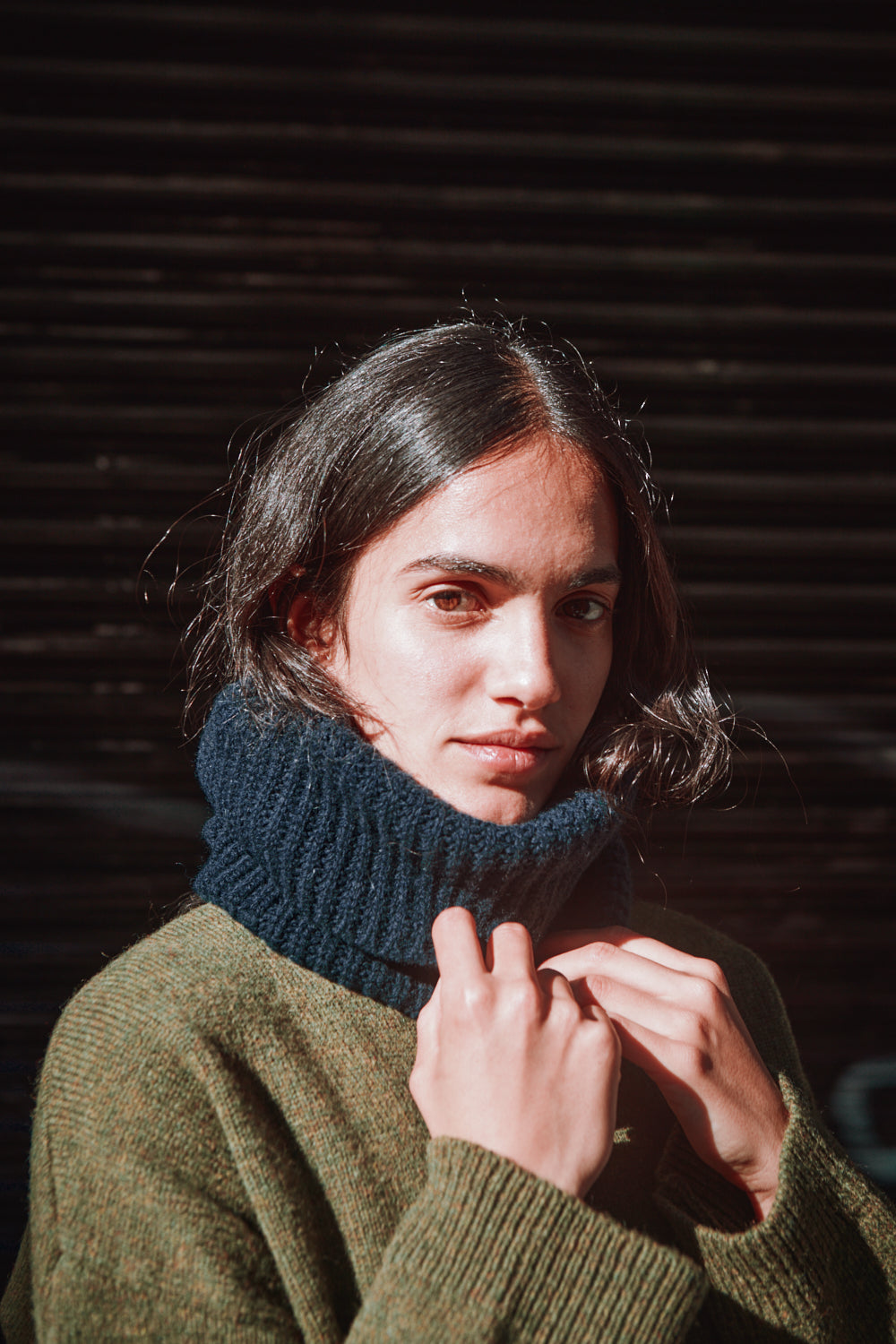 Woman wearing a green sweater and blue knitted turtleneck against a dark background