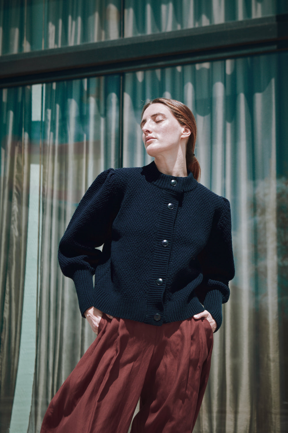 Woman wearing a dark blue  L'ENVERS cardigan and red skirt standing in front of a glass building.