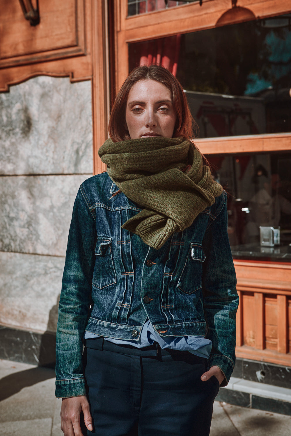Woman wearing a green scarf and denim jacket standing in front of a wooden door.