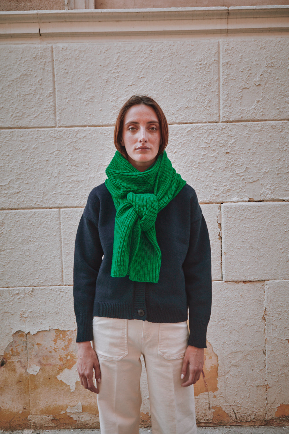 Person wearing a green scarf and dark sweater against a textured wall.