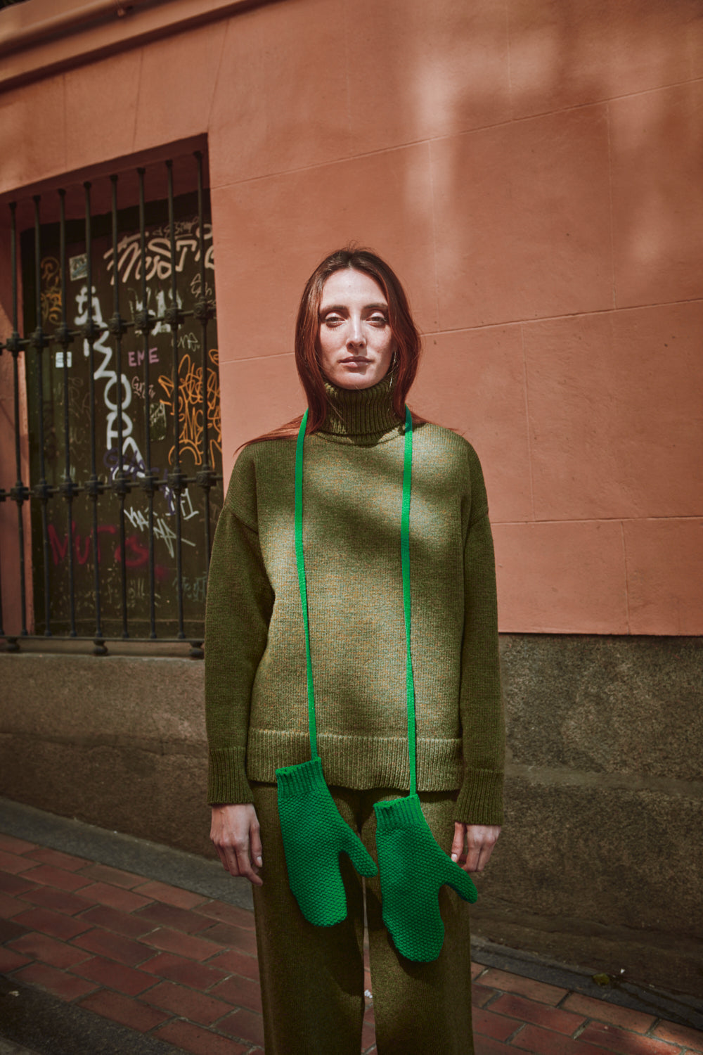 Person wearing a green sweater and matching mittens standing against a wall with graffiti.