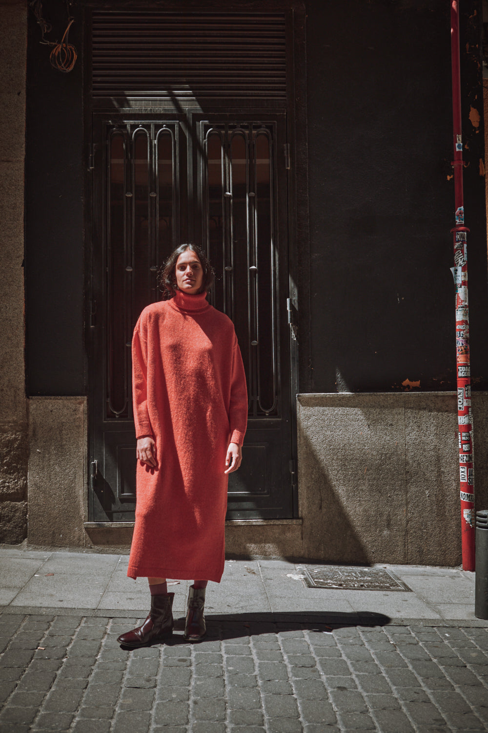 Person wearing a long coral dress standing on a street corner with a dark door in the background.
