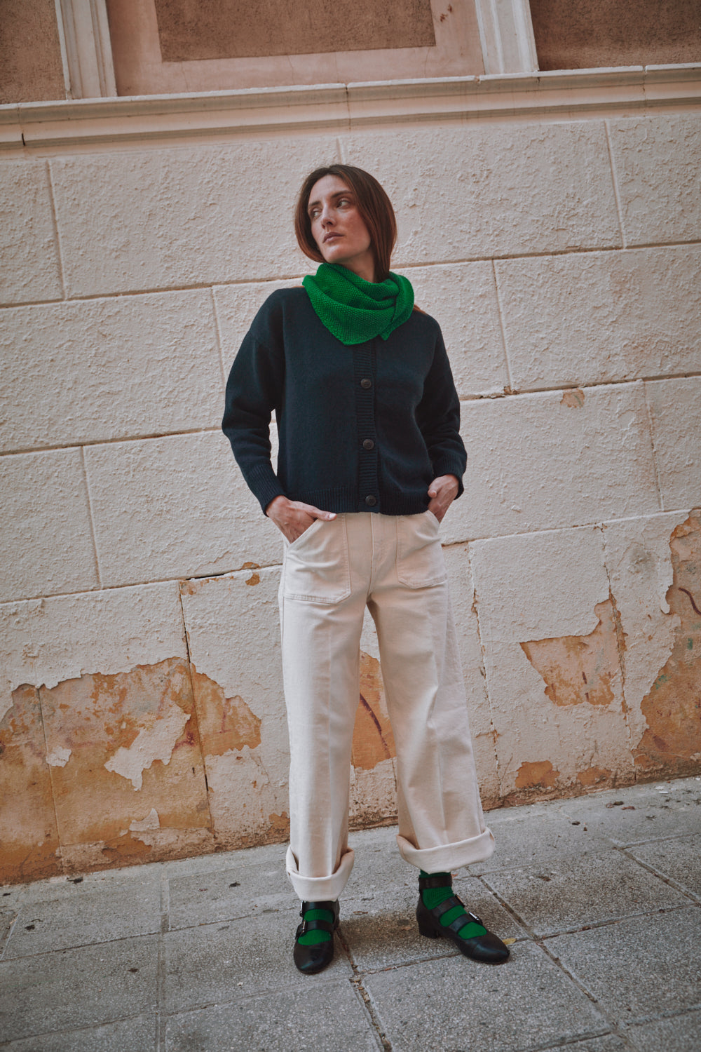 Person wearing a dark sweater, green scarf, and white pants standing against a textured wall.