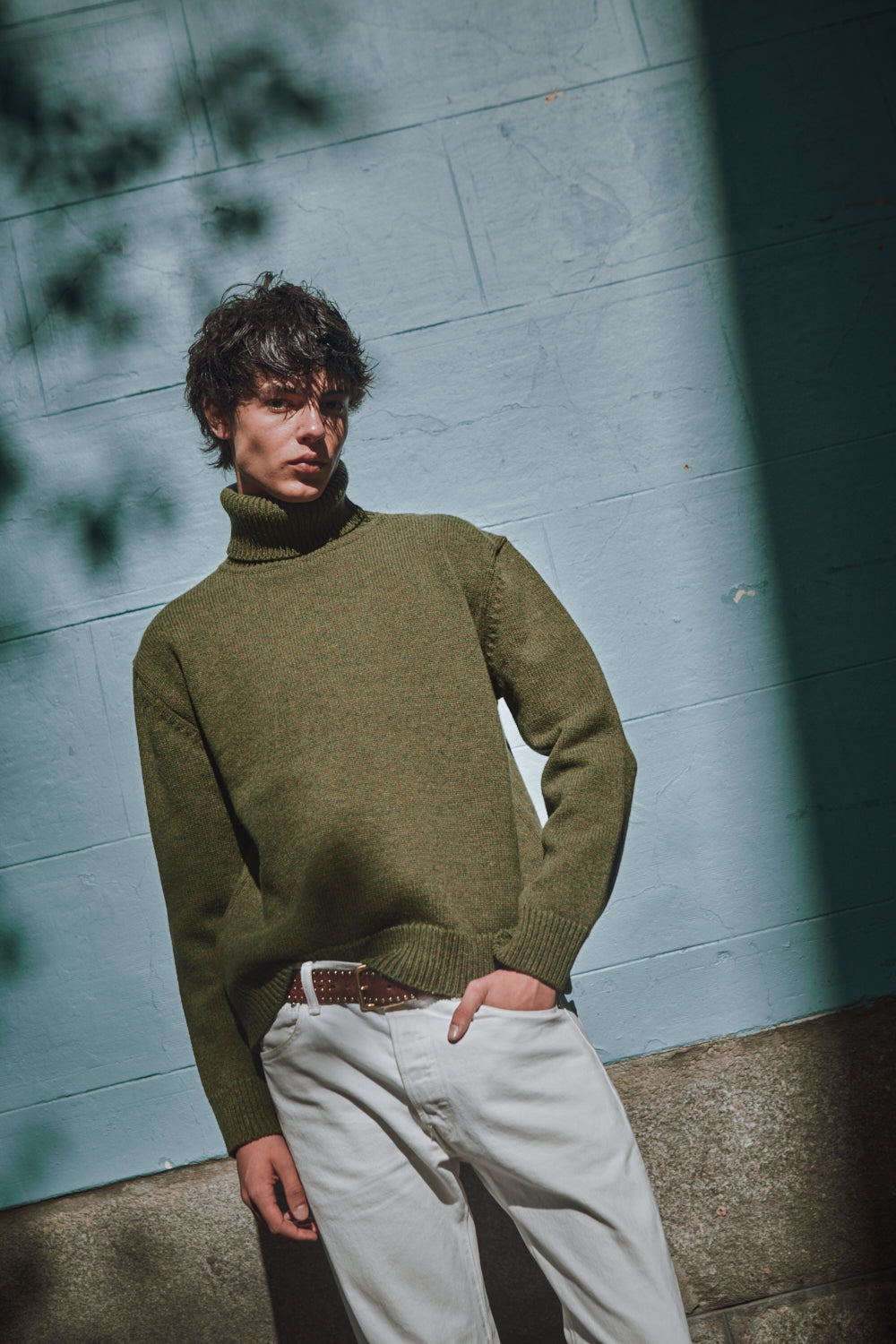Man wearing a green sweater and light-colored pants against a textured wall.