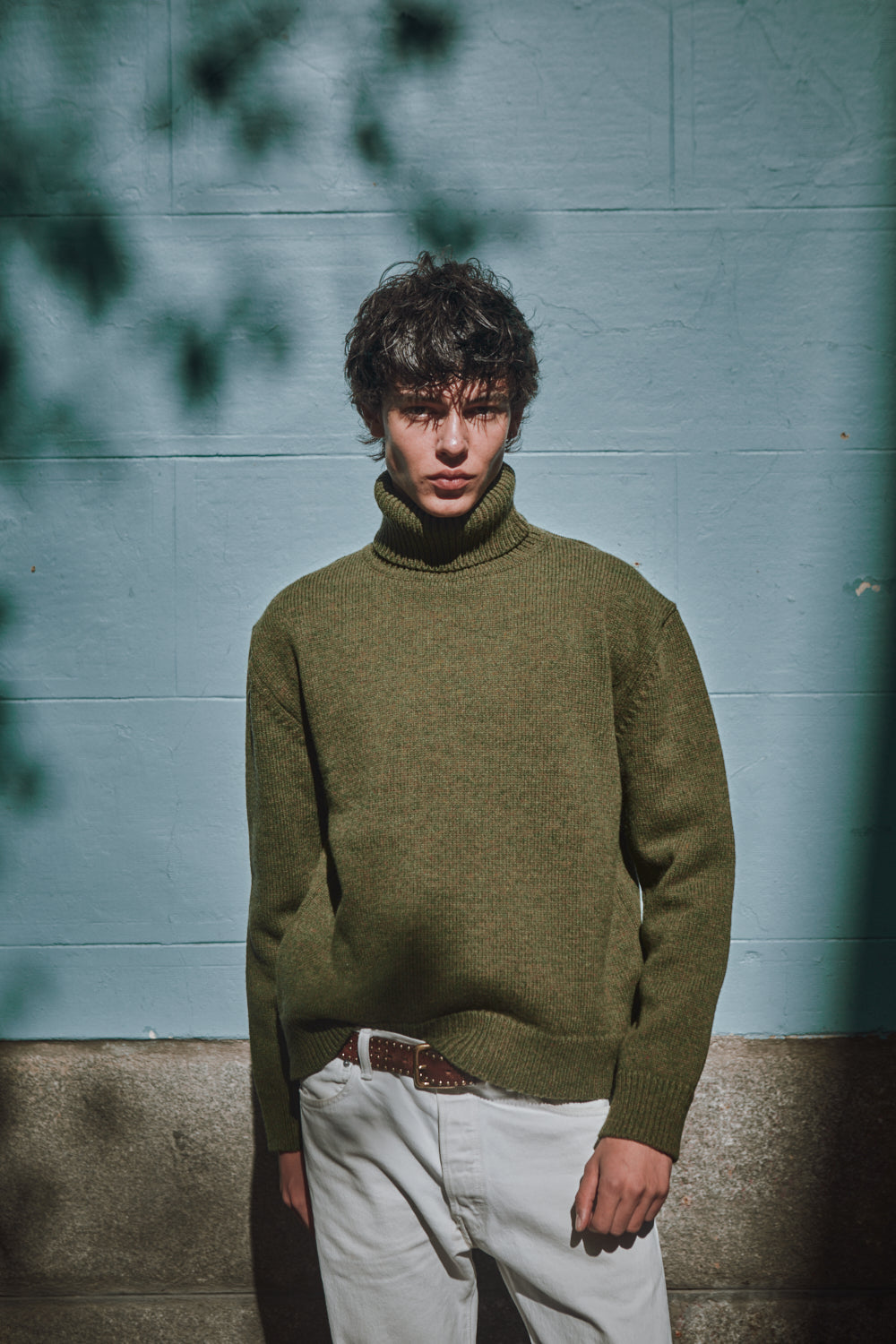 Man wearing a green sweater and light-colored pants against a textured wall.