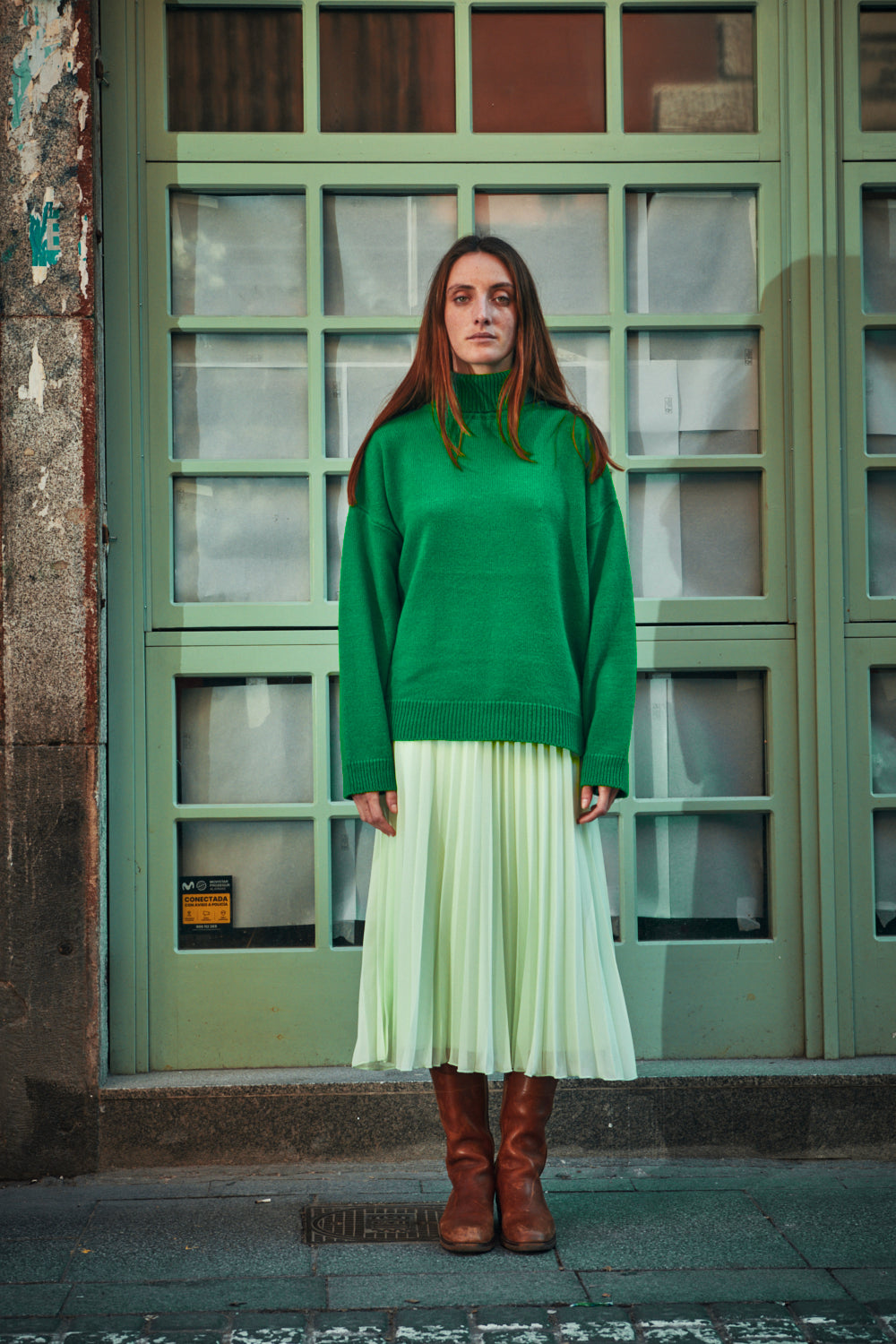 Woman wearing a green L'ENVERS sweater and light green skirt standing in front of a green door.