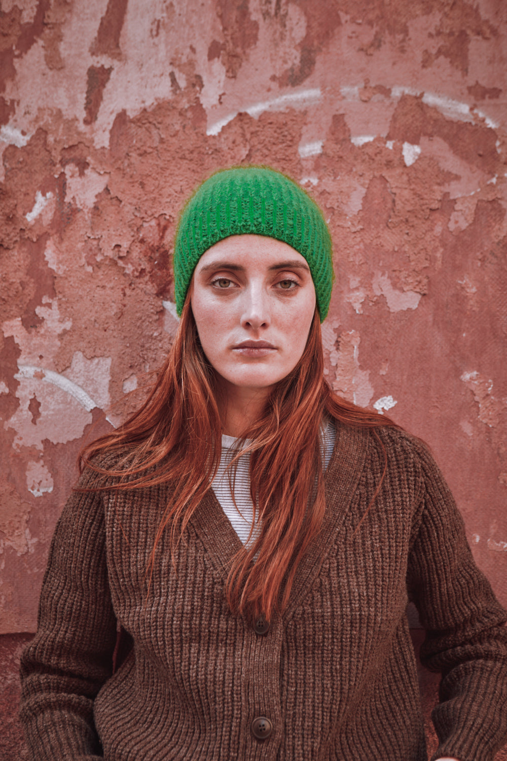 Person wearing a green beanie and brown sweater against a textured pink wall
