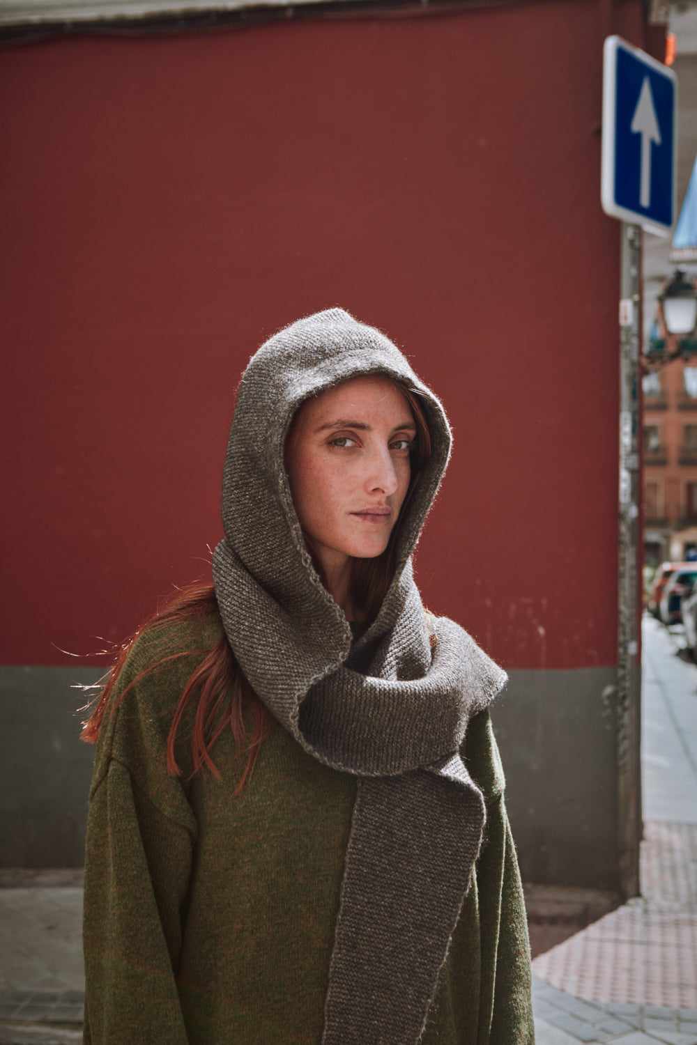 Woman wearing a green coat and gray hooded scarf against a red wall.