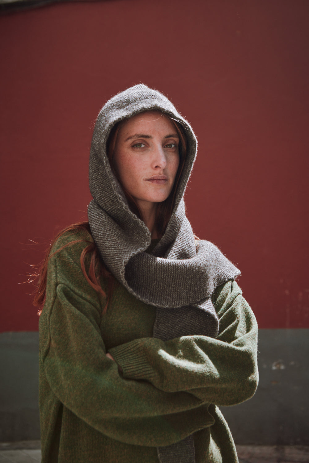 Person wearing a green coat and gray hood against a red wall