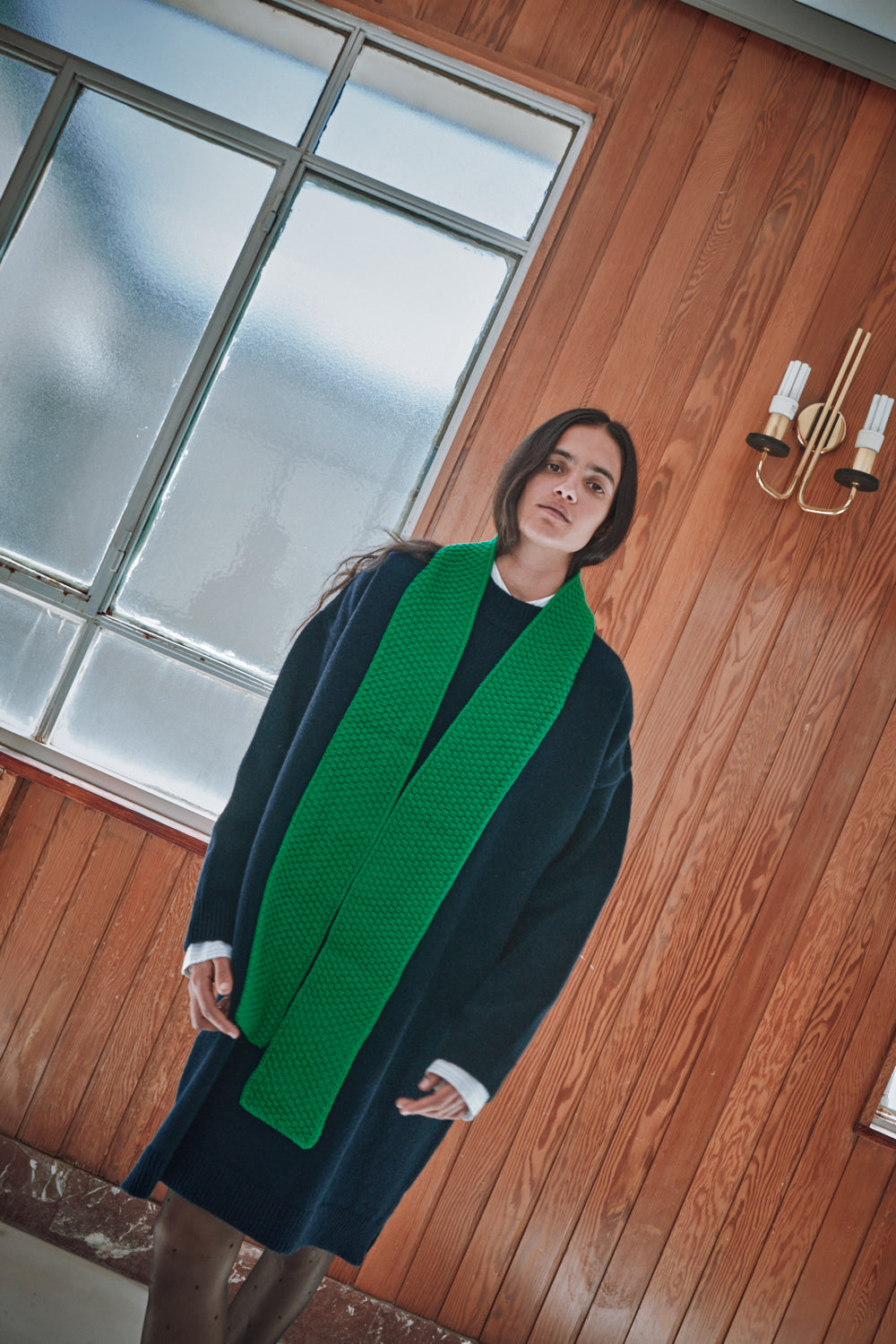 Person wearing a green scarf and dark coat indoors with wooden walls and a window.