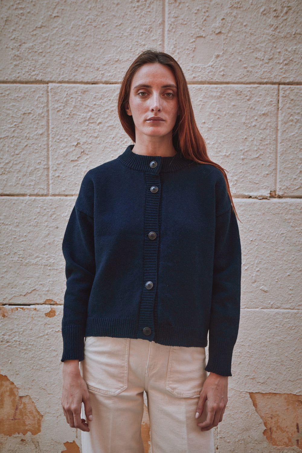 Woman wearing a L'ENVERS navy blue cardigan standing against a textured stone wall