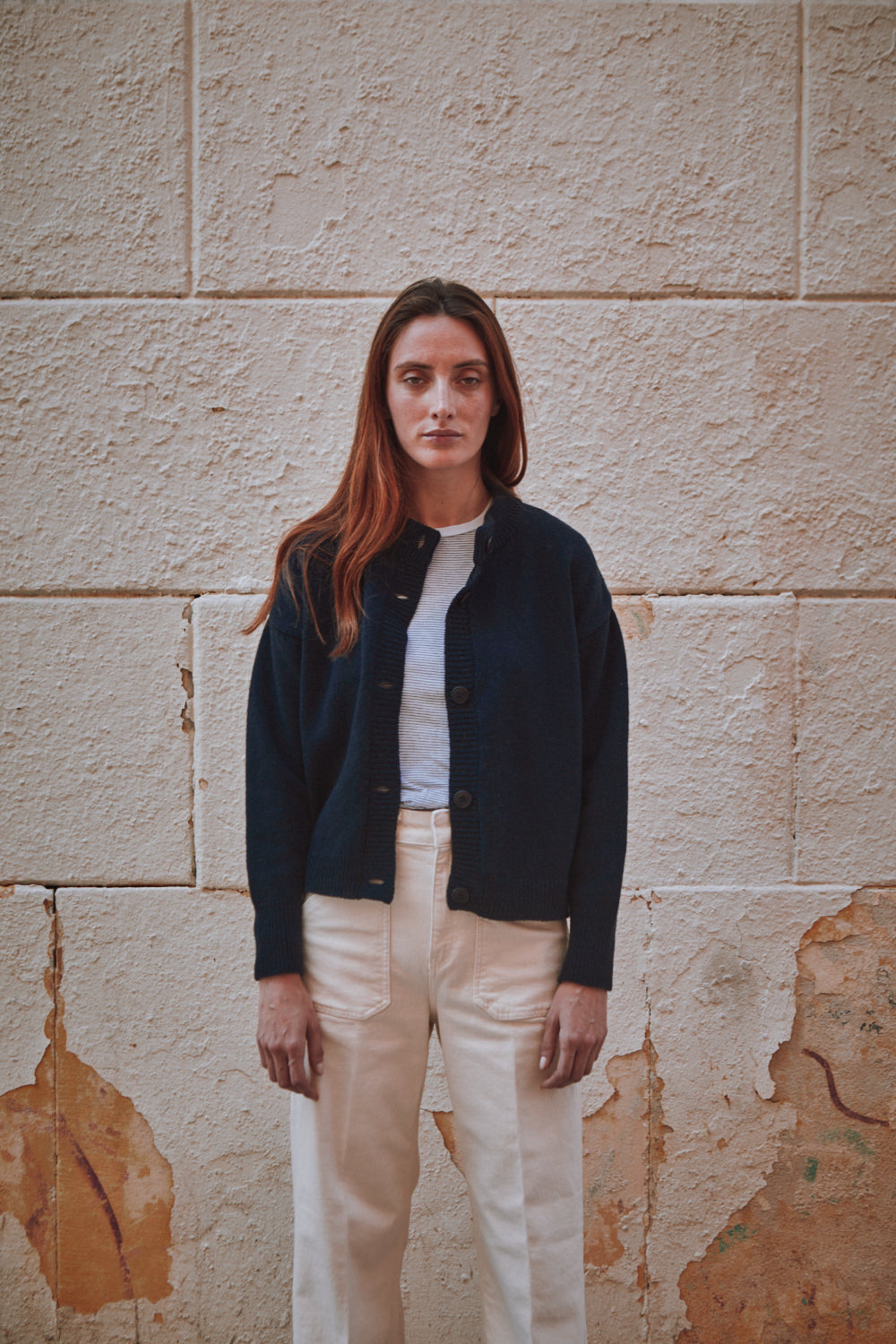Woman wearing a L'ENVERS navy blue cardigan standing against a textured stone wall