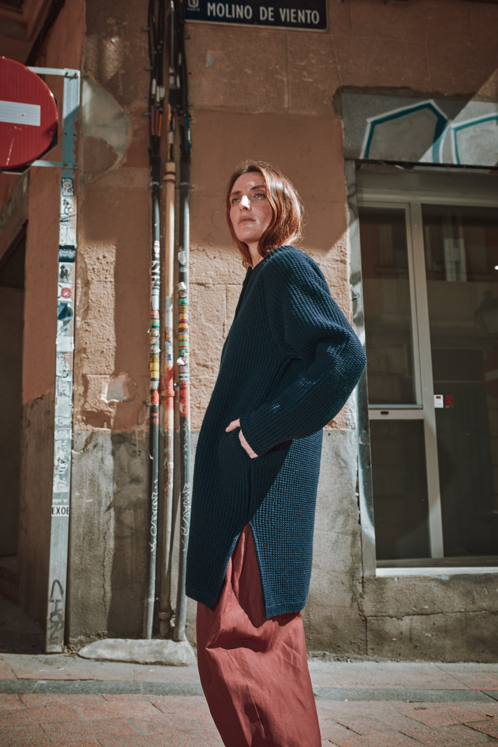 Personne portant une veste bleu foncé et un pantalon rouge, debout au coin d'une rue.