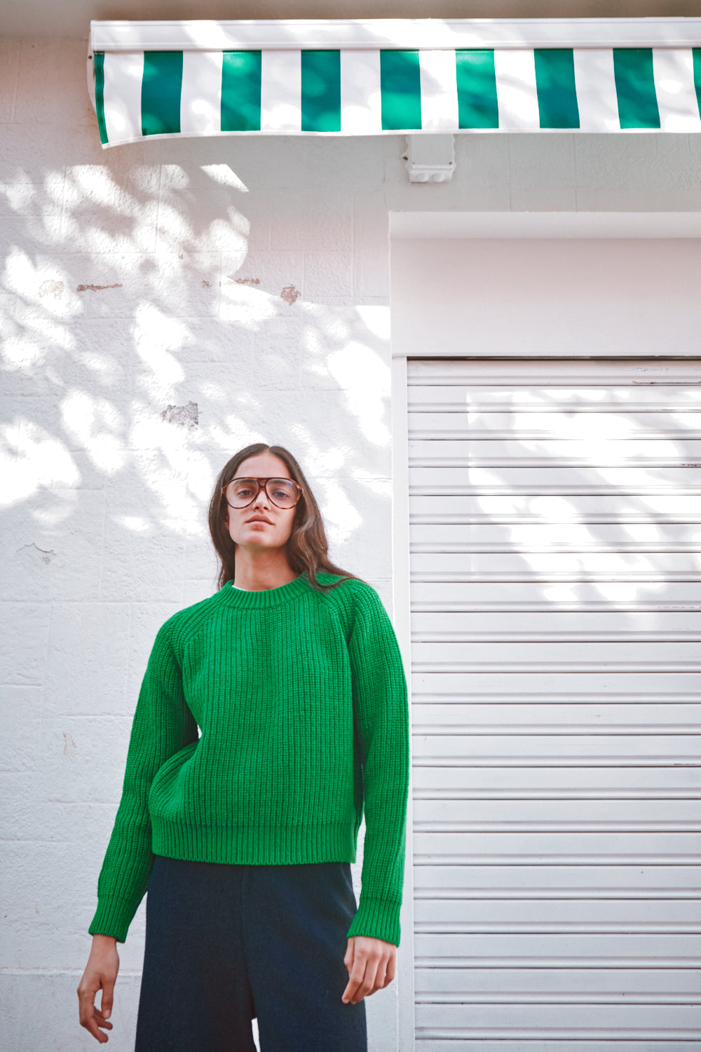 Personne portant un pull vert devant un mur blanc surmonté d'un auvent rayé vert et blanc.