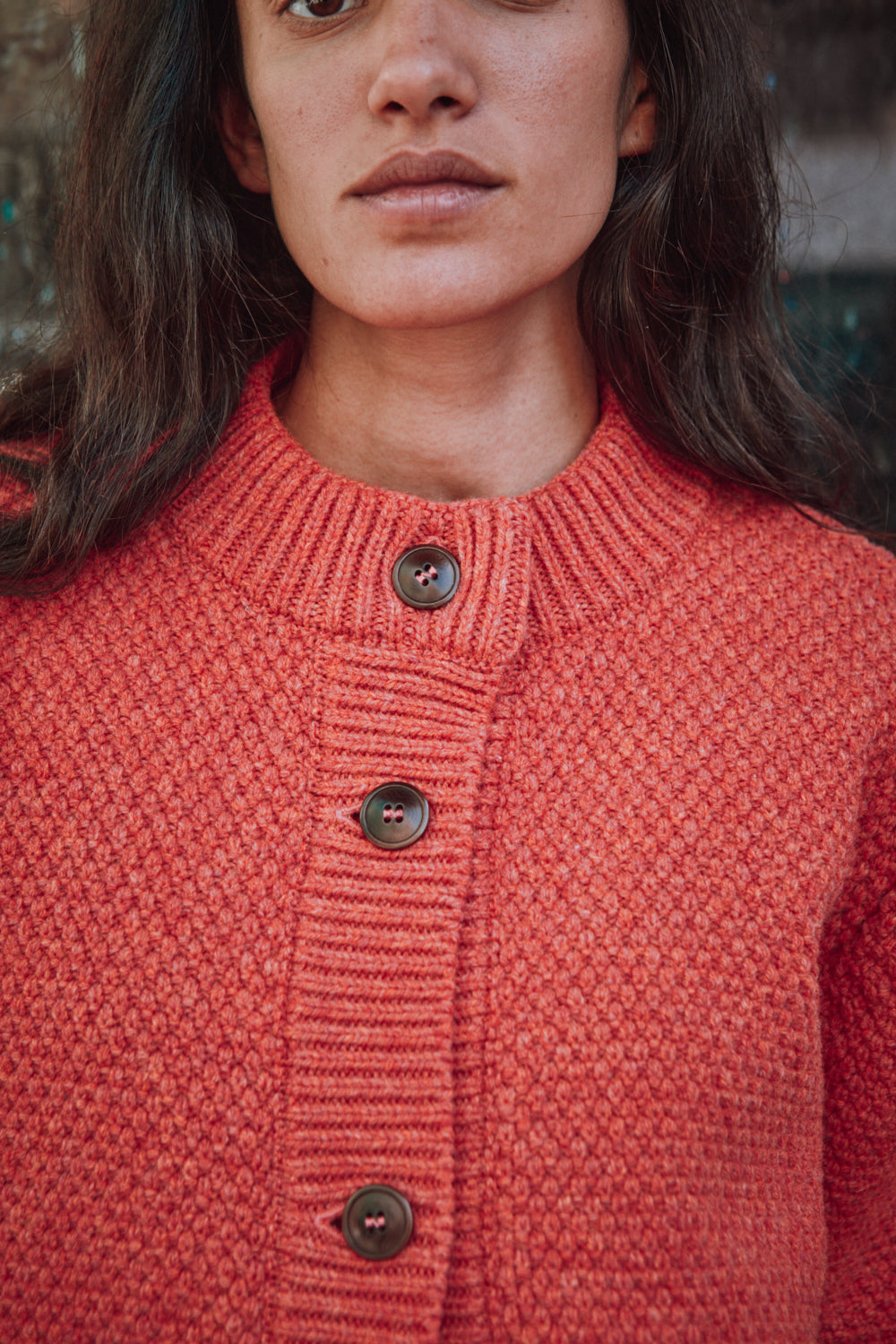 Personne portant un gilet L'ENVERS en tricot rouge devant un mur texturé.