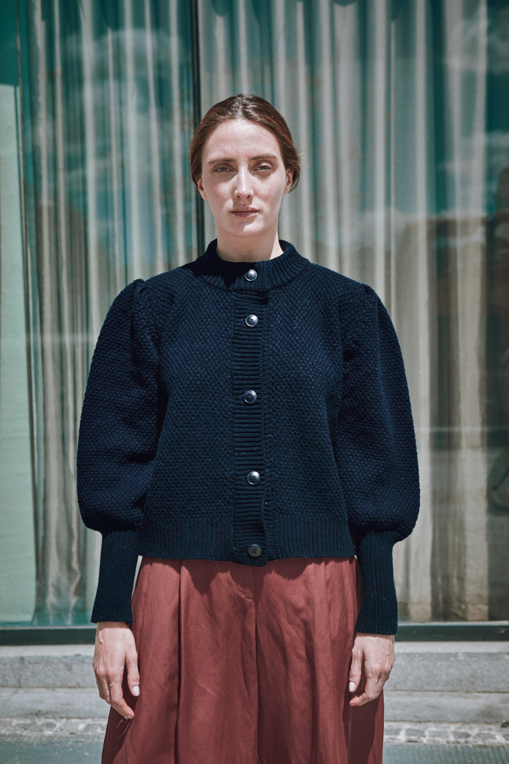 Femme portant un gilet bleu foncé et une jupe rouge de L'ENVERS devant un bâtiment en verre.