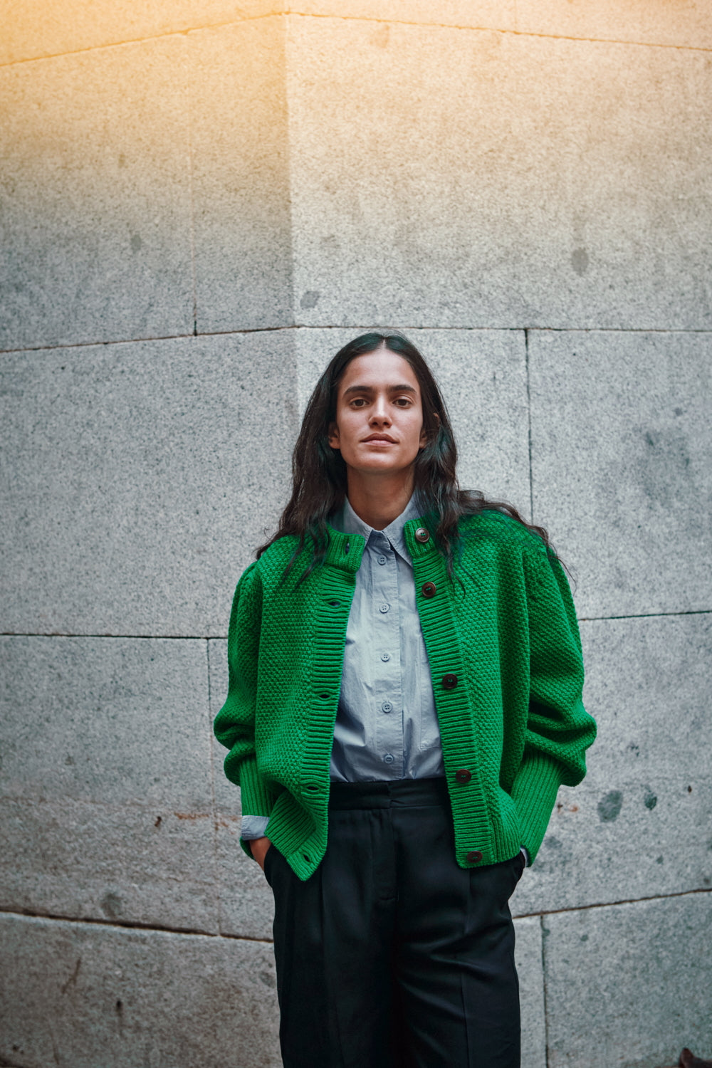 Femme portant un pull en tricot vert de L'ENVERS contre un mur texturé.