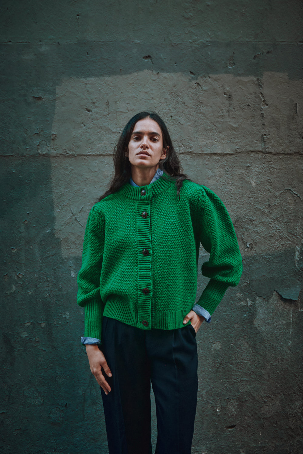 Femme portant un pull en tricot vert de L'ENVERS contre un mur texturé.
