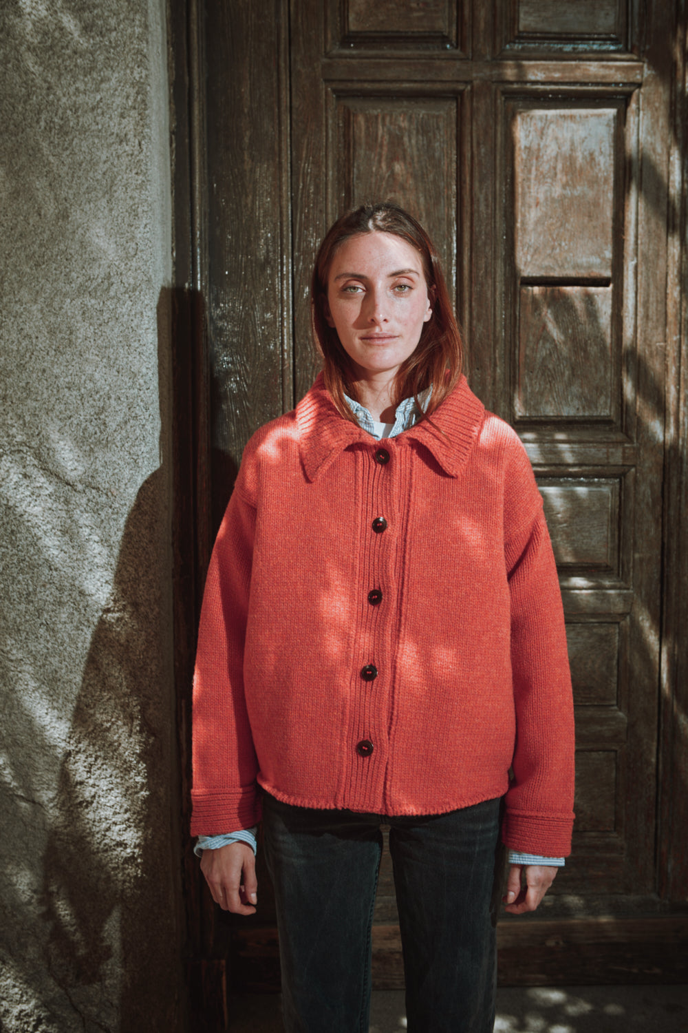 Personne portant une veste corail de L'ENVERS devant un mur texturé et une porte en bois.