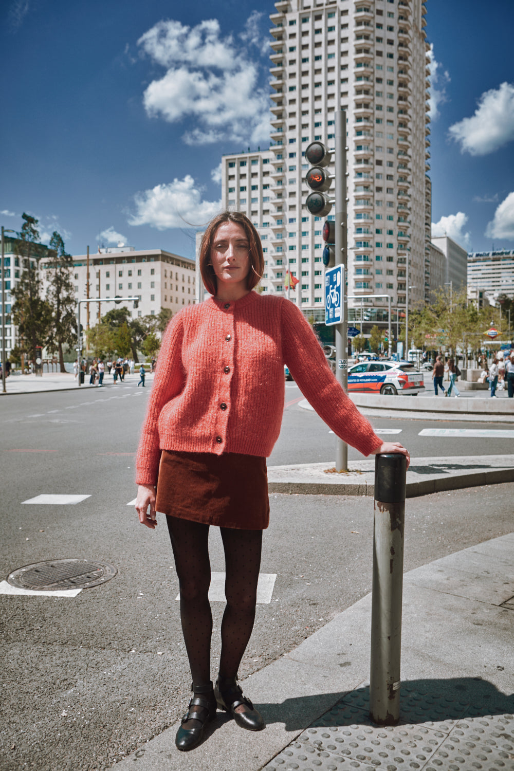 Femme en pull et jupe rose debout dans une rue de la ville avec de grands immeubles en arrière-plan.