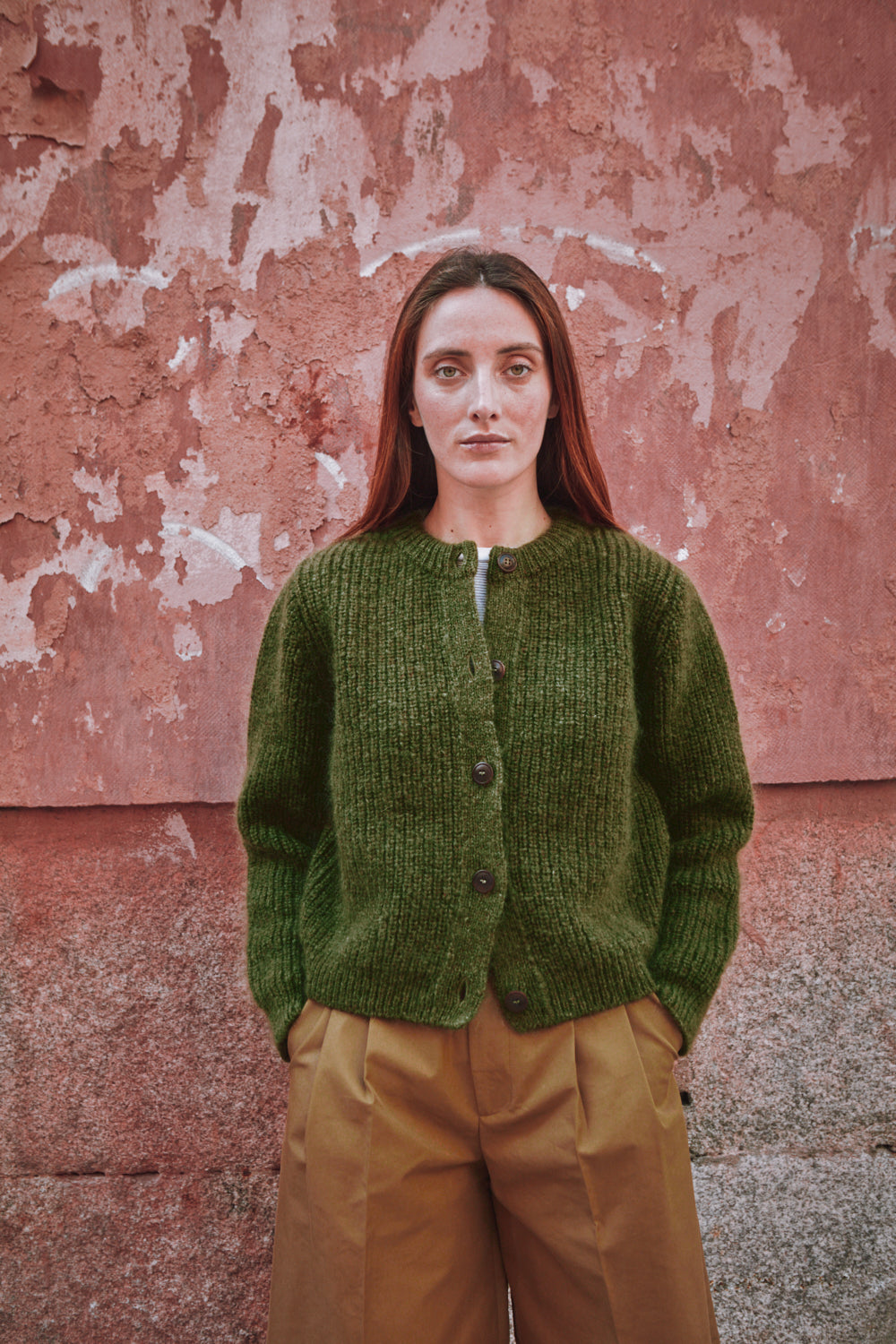 Femme portant un gilet en tricot vert sur un mur rose texturé.