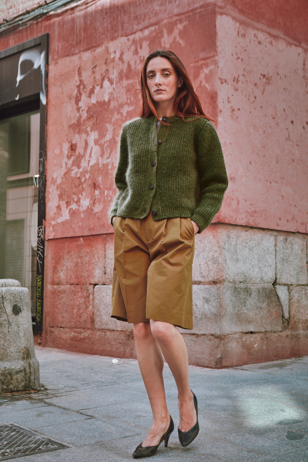 Femme portant un gilet en tricot vert et un short marron, debout contre un mur rouge et gris.