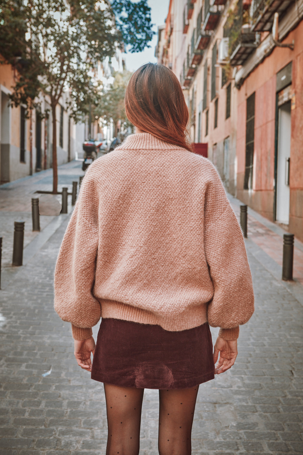 Personne portant un pull rose et une jupe marron marchant dans une rue.