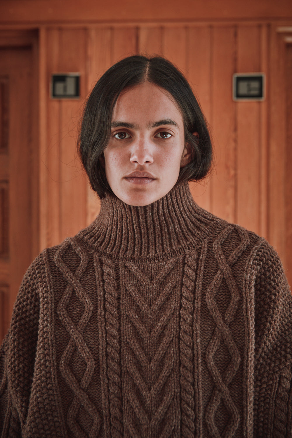 Personne portant un pull en tricot marron de L'ENVERS, debout devant un panneau en bois.