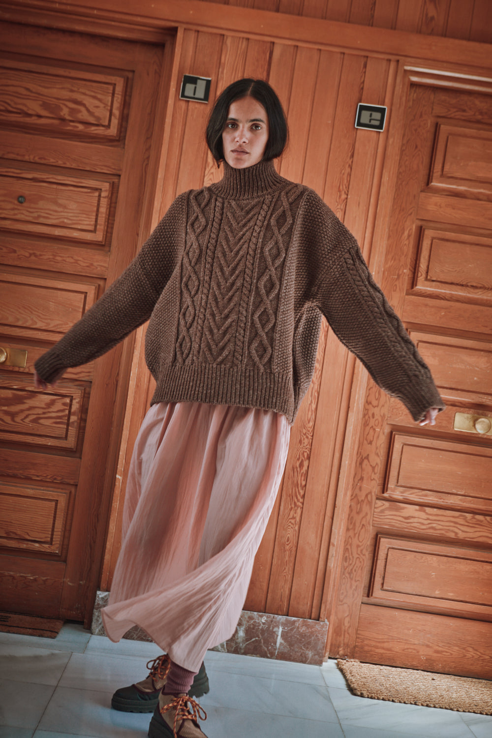 Personne portant un pull en maille marron de L'ENVERS et une jupe rose, debout devant un panneau en bois.
