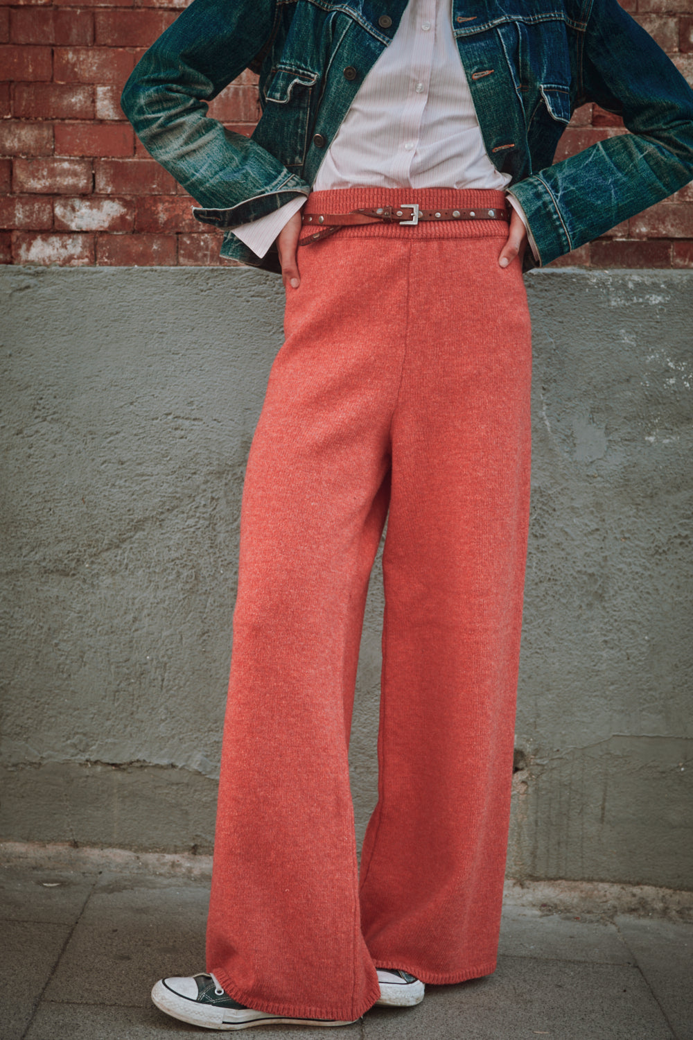 Personne portant une veste en jean, une chemise blanche et un pantalon large rouge contre un mur de briques.
