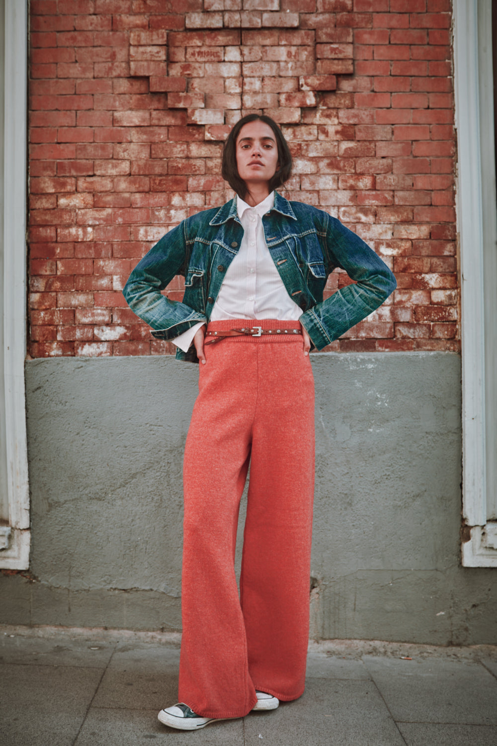 Personne portant une veste en jean, une chemise blanche et un pantalon rouge, debout contre un mur de briques.