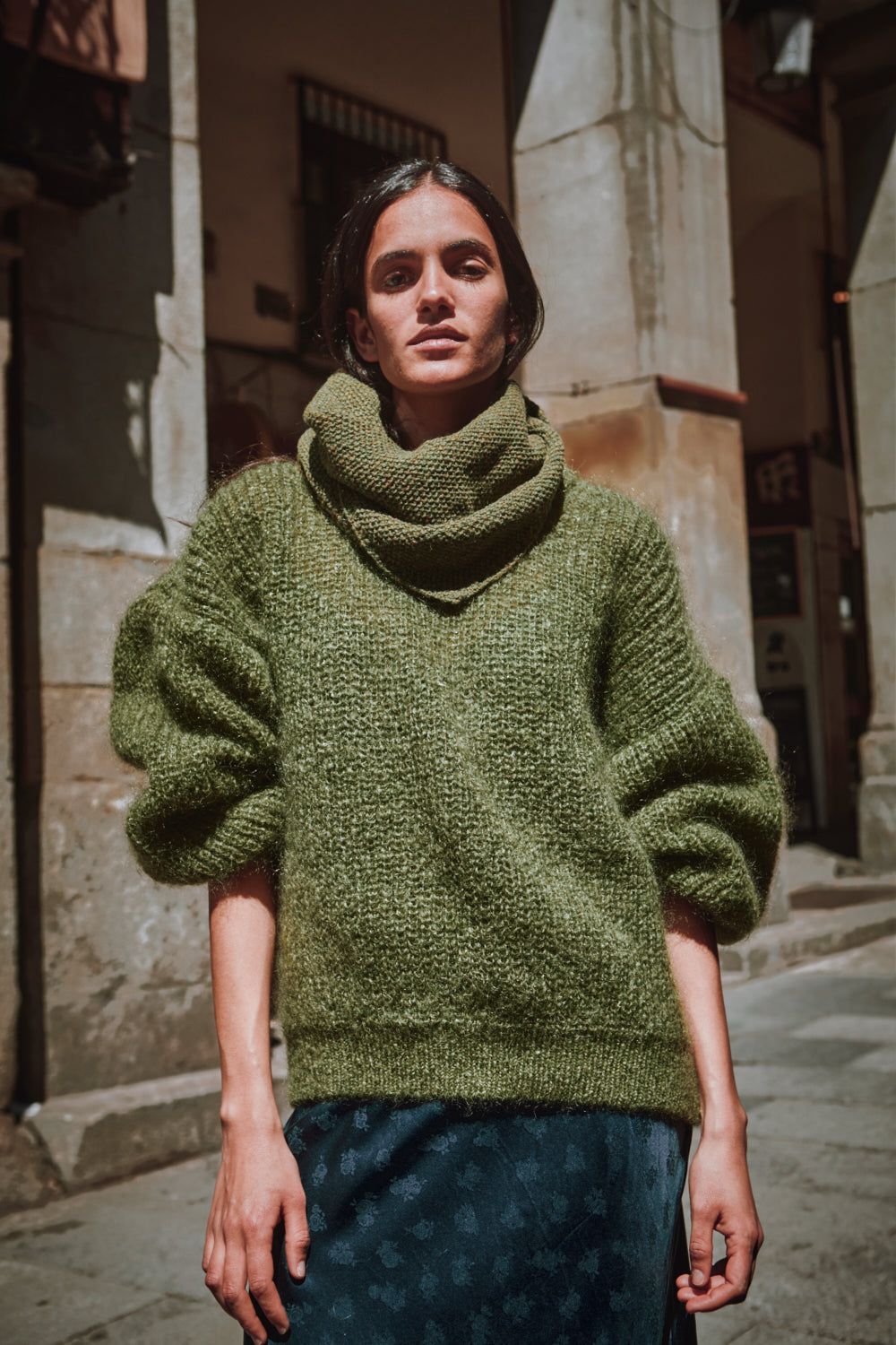 Personne portant un pull vert tricoté, debout dans une rue.