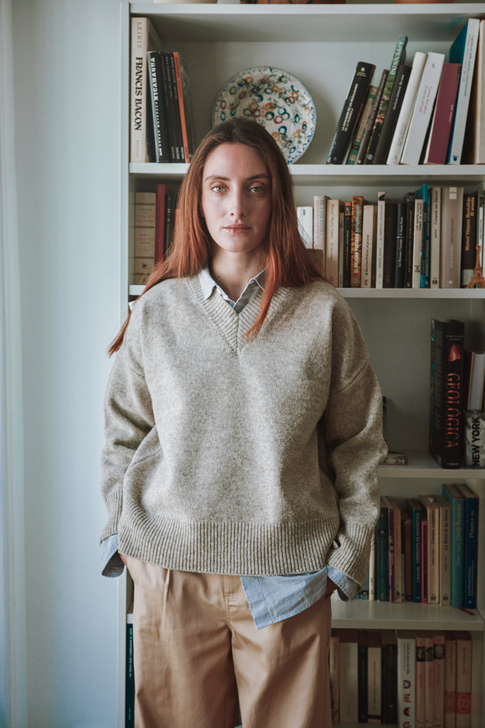 Personne portant un pull beige et un pantalon marron, debout devant une étagère.