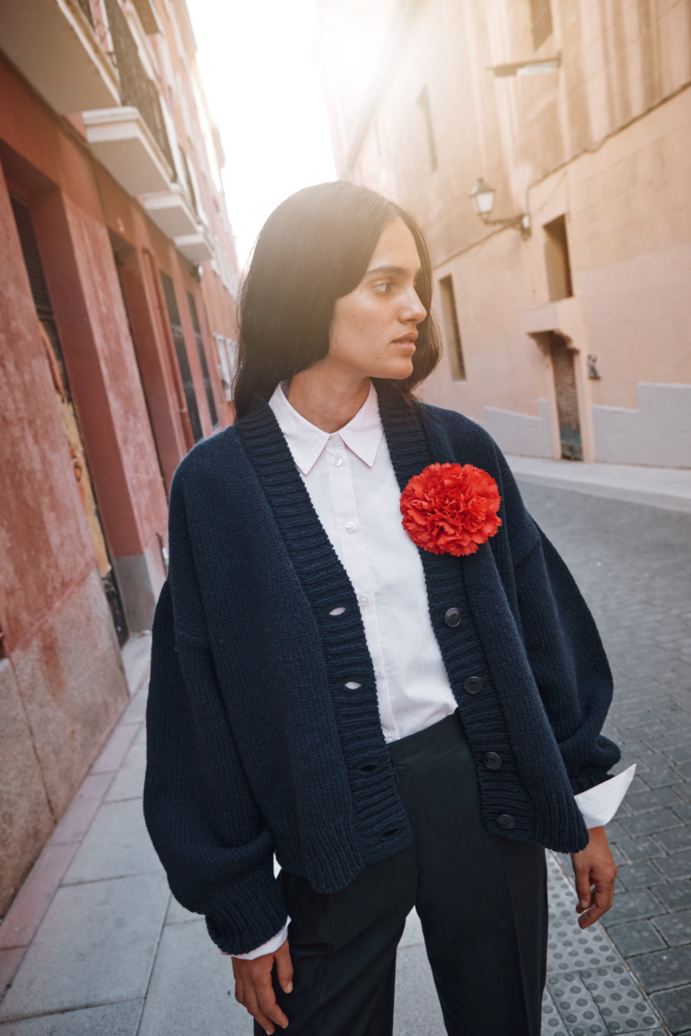 Femme portant un gilet bleu marine sur une chemise blanche avec un accessoire à fleurs rouges, debout dans une rue.