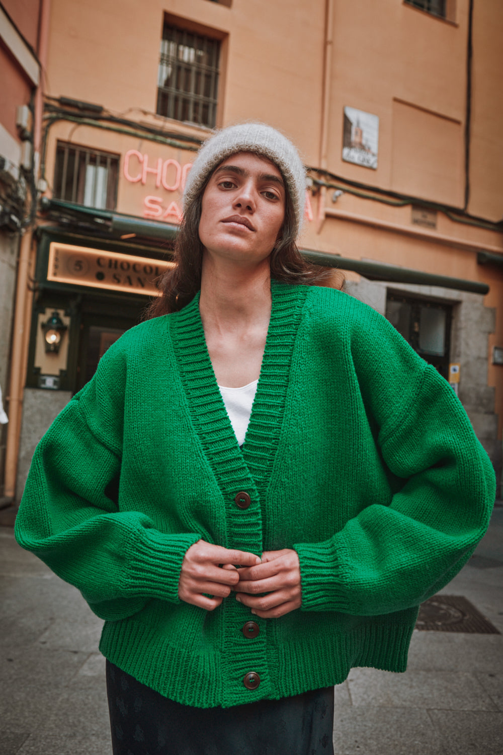 Femme portant un gilet vert et un bonnet blanc, debout dans une rue avec un bâtiment en arrière-plan.